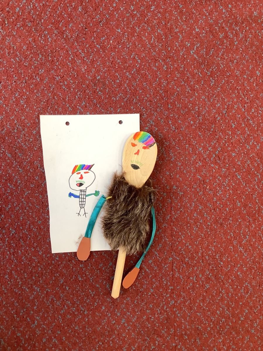 Wooden spoon with face with rainbow hair, fur coat and blue arms next to drawing of the same